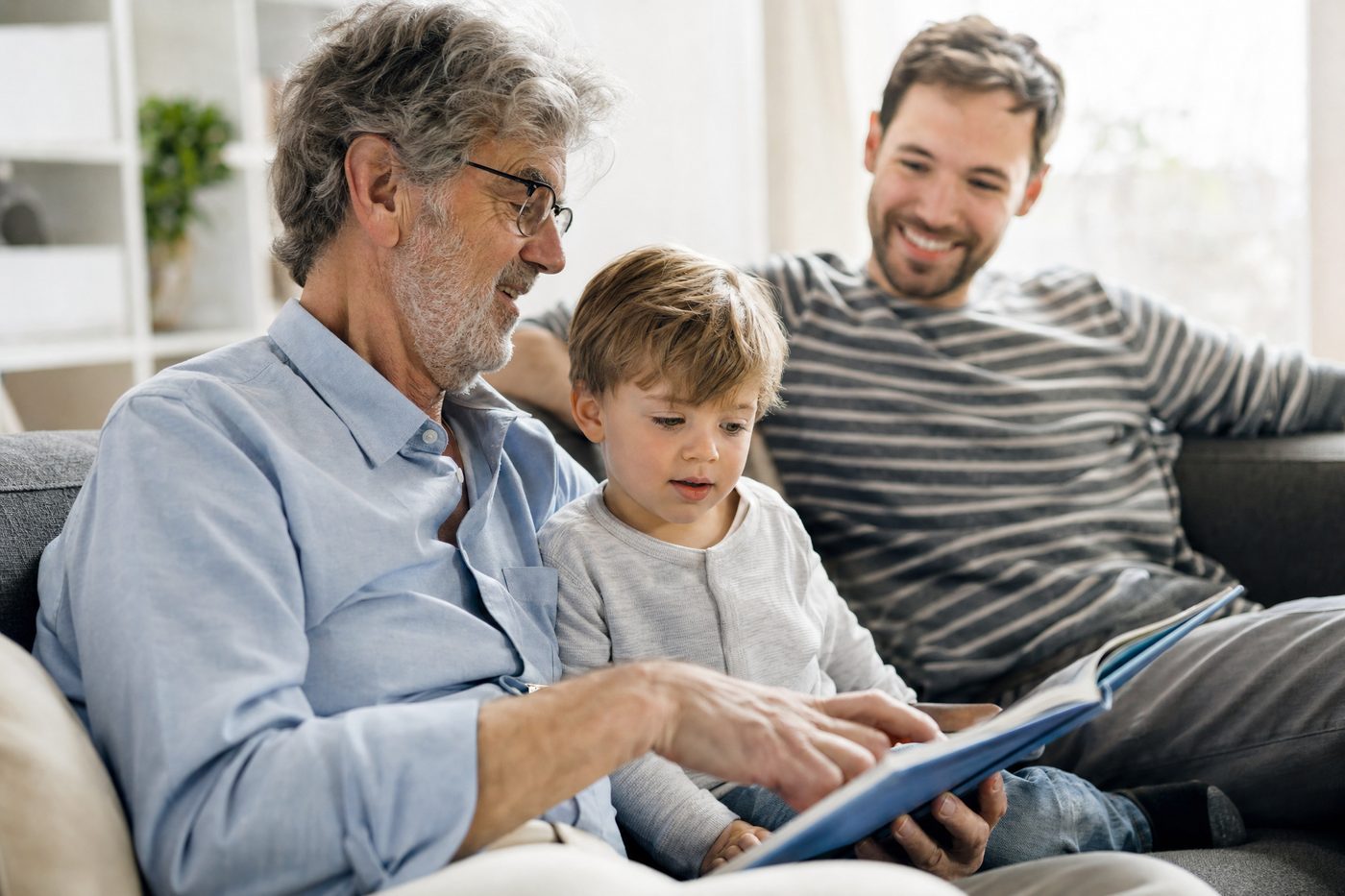 A grandfather reading a book with his young grandson on a sofa while the boy's dad smiles beside them