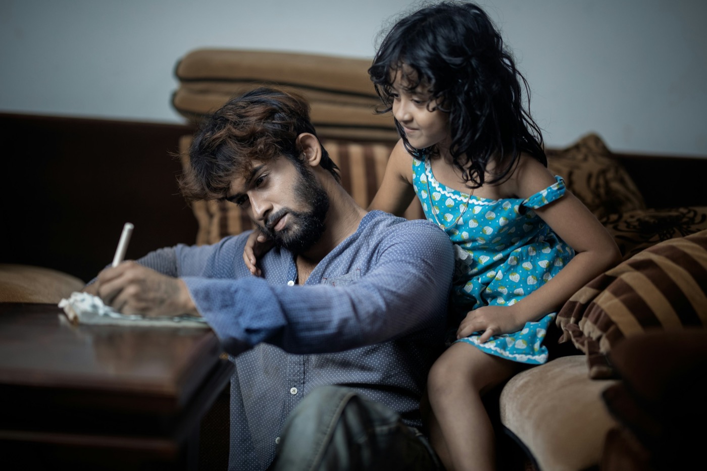 A dad working through paperwork at a table while his daughter leans over his shoulder