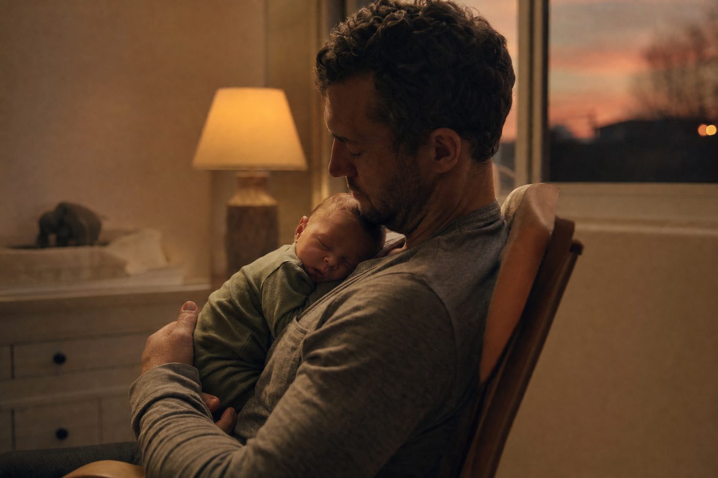 A dad holding his sleeping newborn on his chest in a dim nursery at dusk